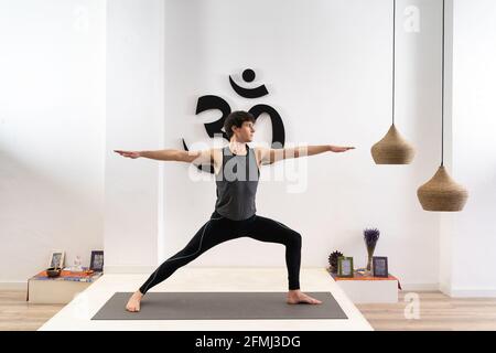 Graceful young male standing in Virabhadrasana and practicing yoga on mat in studio Stock Photo