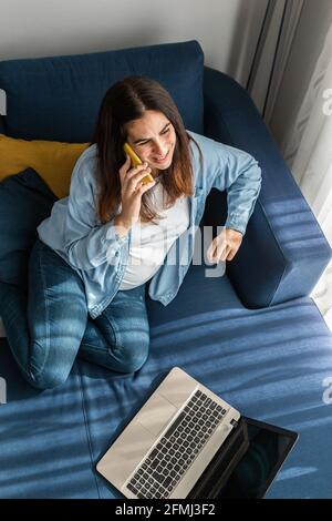 Pregnant female freelancer sitting on sofa with laptop in living room and speaking on mobile phone while working from home Stock Photo