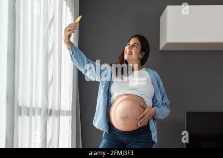 Smiling pregnant female touching belly while standing in room at home and taking selfie with mobile phone Stock Photo