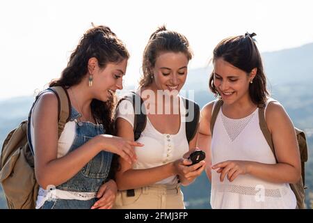 Happy young female travelers in summer clothes using compass together while standing on lush sunny hilly terrain Stock Photo