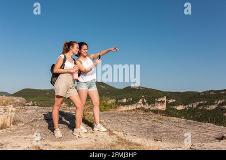 Happy young female travelers in summer clothes using compass together while standing on lush sunny hilly terrain Stock Photo