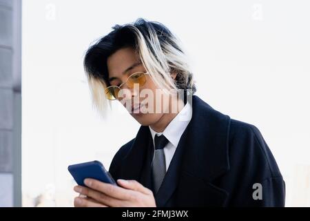 Young well dressed ethnic male entrepreneur in eyeglasses with dyed hair surfing internet on cellphone Stock Photo