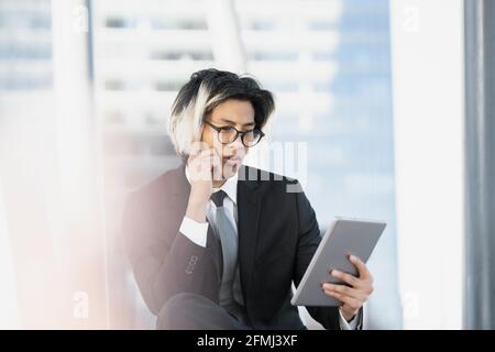 Young well dressed ethnic thoughtful male executive browsing internet on tablet while sitting looking away on city street Stock Photo