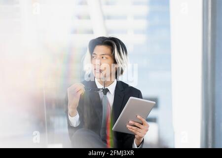 Young well dressed ethnic thoughtful male executive browsing internet on tablet while sitting looking away on city street Stock Photo