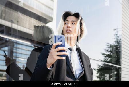 Young Asian male executive in formal apparel text messaging on mobile phone in daylight looking away Stock Photo
