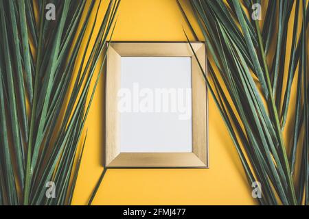 Wooden frame with white picture hanging on orange wall decorated with fresh green palm leaves at home Stock Photo