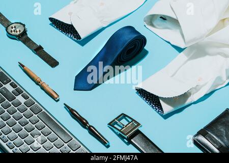 From above of elegant white shirt with blue silk tie and wristwatch arranged on table with leather belt and creative pens near computer keyboard Stock Photo