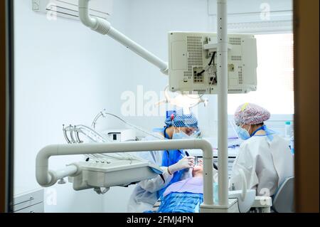 Anonymous dentist with colleague in uniforms operating teeth of patient using medical tools in modern clinic Stock Photo