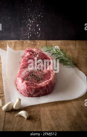 Raw beef piece with ground black pepper under salt in air between fresh garlic cloves and rosemary sprig Stock Photo