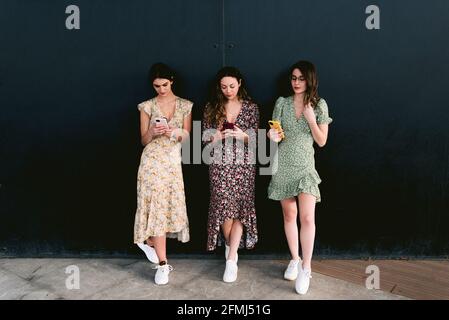 Young best female friends in trendy apparel with cellphones standing on urban walkway against wall Stock Photo