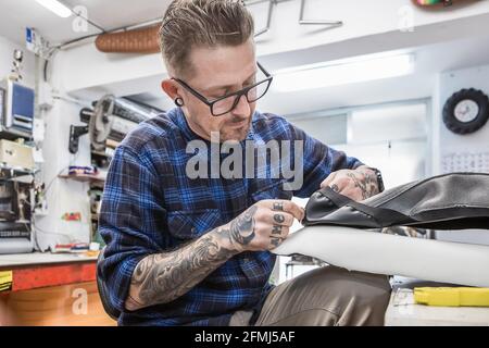 Creative bearded craftsman sewing leather upholstery for motorbike seat while working in workshop Stock Photo