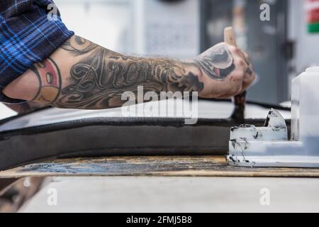 Crop side view of tattooed craftsman applying glue on motorcycle seat while making upholstery at workbench Stock Photo