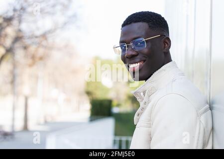 Side view of young cheerful confident black male entrepreneur with stylish jacket looking at camera standing in the city Stock Photo