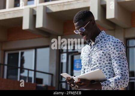 Content African American businessman in ornamental shirt chatting on cellphone while standing with tablet in town Stock Photo