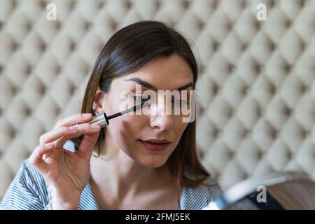 Focused young brunette in blue dress applying mascara on eyelashes and looking in round mirror at home Stock Photo