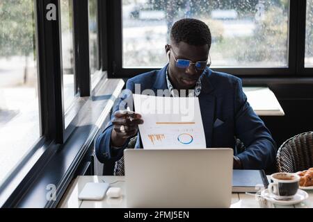 Young ethnic male entrepreneur in eyeglasses with graphic on paper working at cafe table Stock Photo