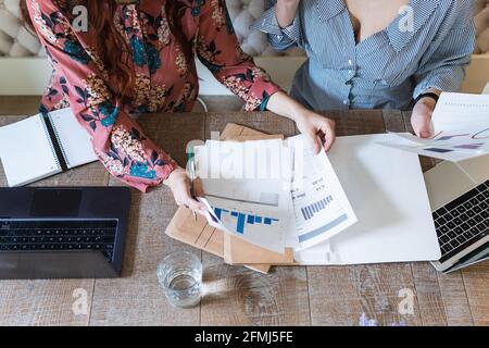 From above crop female coworkers gathering at desk with laptops and analyzing diagrams while working together in modern workspace Stock Photo