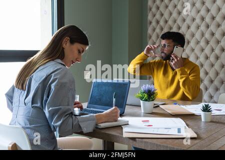 Concentrated young multiracial coworkers in casual wear working on netbooks taking notes and talking on smartphone while working together in modern li Stock Photo