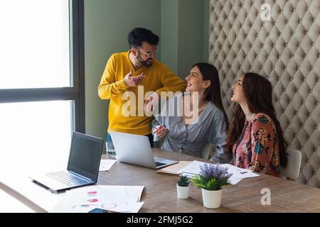 Group of happy laughing coworkers in casual clothes gathering at desk with laptops and discussing business details while working together in light con Stock Photo