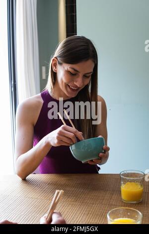 Happy young female wearing casual outfit enjoying tasty food with chopsticks while sitting at table in modern dining room Stock Photo