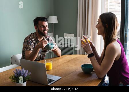 Cheerful young friends wearing casual outfits enjoying tasty lunch and chatting while sitting at dining table with opened netbook in modern apartment Stock Photo