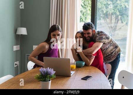 Group of young cheerful friends wearing casual outfits embracing and gathering at dining table while browsing modern netbook and smiling Stock Photo