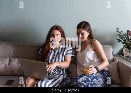 Content best female friends with beverage surfing internet on netbook while sitting on sofa in house Stock Photo