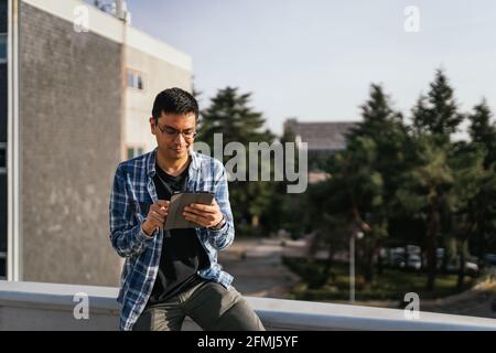 Concentrated male wearing eyeglasses and checkered shirt browsing modern tablet while standing on rooftop in suburb on sunny summer day Stock Photo