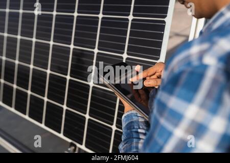 Cropped unrecognizable ethnic male technician in checkered shirt browsing tablet while standing near photovoltaic panel located in modern solar power Stock Photo