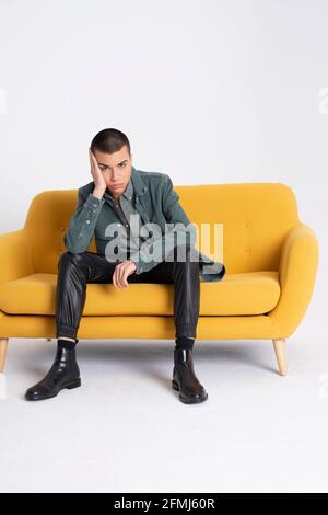Young handsome male model in trendy clothes sitting on yellow couch on white background in studio Stock Photo