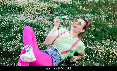 Expressive young female in bright colorful clothes playing with soap bubbles and sitting on verdant green meadow in nature Stock Photo