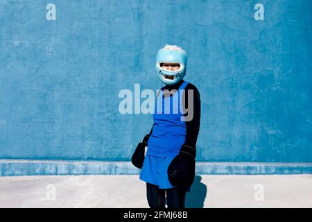 Mature female in sportswear and boxing gloves standing with helmet against blue wall and looking at camera Stock Photo
