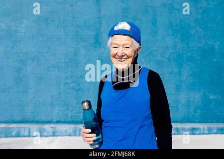 Cheerful mature sportsWoman in activewear and cap standing with water bottle in hand against blue wall in sunny outdoor training center and looking aw Stock Photo