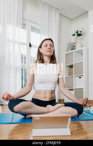 Tranquil female sitting in Lotus pose and listening to meditation on tablet while practicing yoga at home Stock Photo
