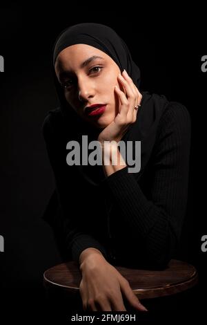 Attractive young Islamic female wearing black outfit and hijab touching face gently leaning on chair in black studio looking at camera Stock Photo
