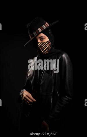 Fashionable Islamic female wearing leather jacket hijab and trendy hat standing looking at camera in dark studio during coronavirus outbreak Stock Photo