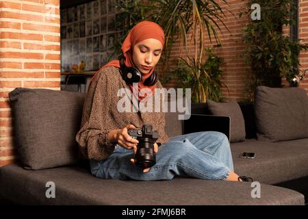 Concentrated young Muslim female in modest wear hijab and headphones using professional photo camera on cozy couch Stock Photo