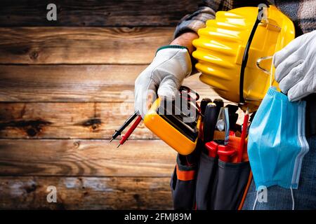 Electrician worker on vintage wooden background; holds the tester, helmet, protective goggles and the surgical mask to prevent the spread of Coronavir Stock Photo