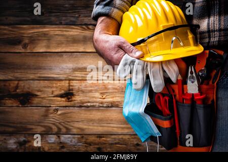 Electrician worker on vintage wooden background; holds helmet, gloves, goggles and the surgical mask to prevent the spread of Coronavirus. Constructio Stock Photo