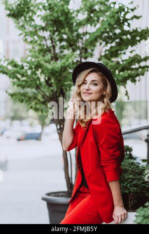Young beautiful blond woman in a red suit and black hat stands outdoors in the yard next to the building. Stylish people. Soft selective focus. Stock Photo