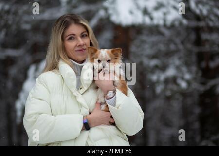 Blonde young female holding ginger and white chihuahua in her hands. Snow and trees on the background. Stock Photo