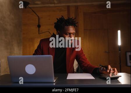 Handsome Black African American Man Working on Laptop Computer while Sitting Behind Desk in Cozy Living Room. Freelancer Working From Home Stock Photo