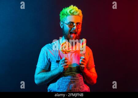Extremely joyful hipster guy in denim overall, holding lollipop and ice cream in hands, screaming with excitement, crazy about sweet sugary confection Stock Photo