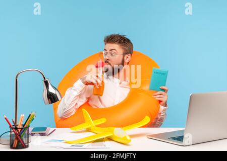 Funny happy man office worker drinking lemonade sitting in rubber lifebuoy and holding passport, going to fly in warm countries, planning vacation. In Stock Photo