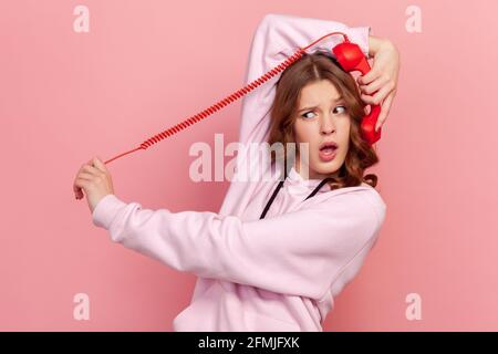 Portrait of amazed curly hair teenage girl holding red telephone handset receiver, fooling around. Indoor studio shot isolated on pink background Stock Photo