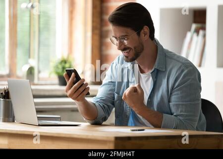 Excited smiling businessman in glasses celebrating success, using phone Stock Photo