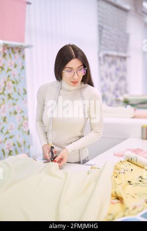 Closeup photo of dressmaker woman hands cutting yellow fabrics with blurred background. Professional instrument concept. Tailor woman cuts linen textile cloth in atelier boutique. High quality photo Stock Photo