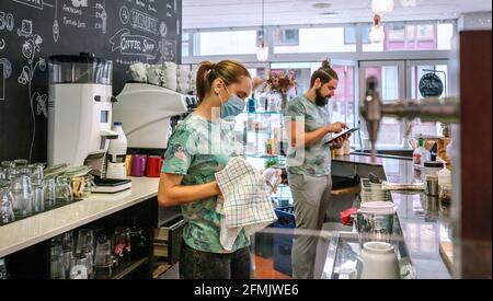 Waitress with mask cleaning glasses while her coworker works with tablet Stock Photo