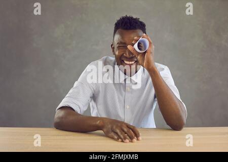 Happy black man sitting at desk and looking through hole in paper roll like in telescope Stock Photo