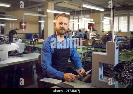 Portrait of a friendly man sitting at his workplace and using a hammer to make belts in a factory. Stock Photo